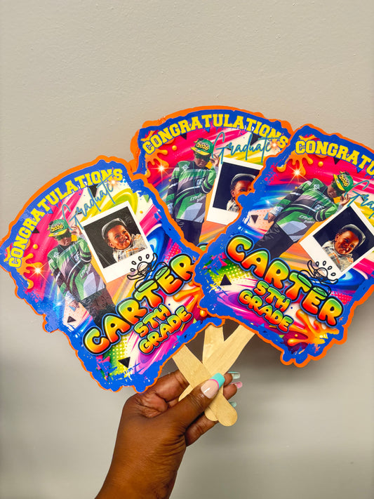 Colorful custom graduation fans with text and images held by a hand against a plain background